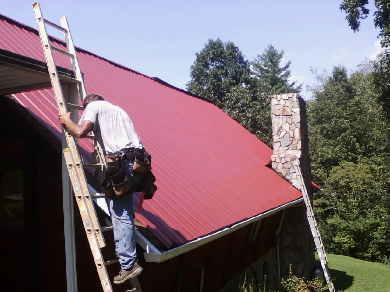Roofer on a ladder beside a red metal roof and stone chimney — Soffit & Fascia Repair by Roofing Brothers in Florence